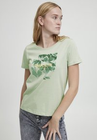 Camiseta de algodón verde claro con cuello redondo y mangas cortas; presenta un estampado botánico en verde y dorado con el texto "Simplicity Made Perfect."