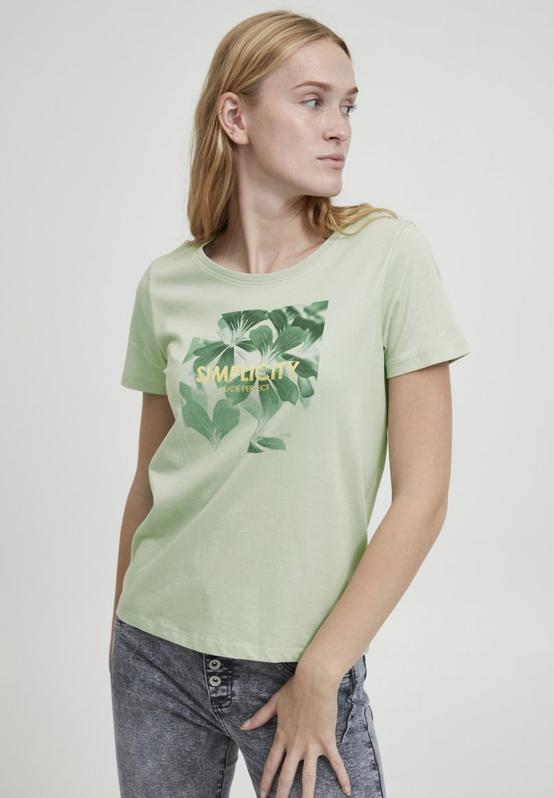 Camiseta de algodón verde claro con cuello redondo y mangas cortas; presenta un estampado botánico en verde y dorado con el texto "Simplicity Made Perfect."
