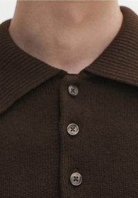 Brązowa dzianinowa koszulka polo z szerokim kołnierzem, z trzema guzikami w dopasowanym kolorze, zbliżenie na teksturę tkaniny.