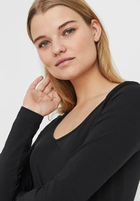 Haut noir à manches longues avec un col rond, texture lisse et design ajusté. Le mannequin a les cheveux blonds lâchés, touchant son visage.