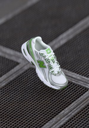 Biały i zielony sportowy sneaker z białymi sznurowadłami, spoczywający na czarnej metalowej powierzchni z siatki.