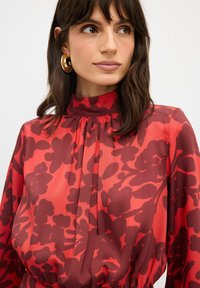 Rode bloes met bloemenprint, hoge hals, geplooide details en lange mouwen. Gemaakt van een gladde, lichte stof met contrasterende donkerrode patronen.