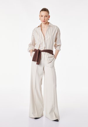 Frau trägt locker sitzendes beiges gestreiftes Hemd mit hochgekrempelten Ärmeln, weit geschnittene cremefarbene Hose, braunen Pullover um die Taille gebunden und schwarze spitze Schuhe.