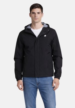 Giovane uomo con giacca nera con cappuccio e logo sul petto, maglietta bianca e jeans blu, in piedi davanti a uno sfondo uniforme.