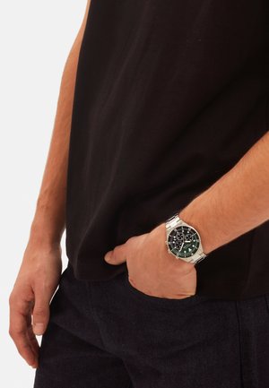 Herrenarmbanduhr mit einem silbernen Edelstahlband, schwarzem Ziffernblatt, grünen Akzenten, Chronographenfunktionen und einem runden Gehäuse. Wird an einer Hand in einer Tasche getragen.