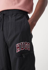 Černé, lehké cargo kalhoty s elastickým pasem a šňůrkou. Obsahují vyšívané logo "HUGO" v červené a bílé barvě a kapsu na zip.