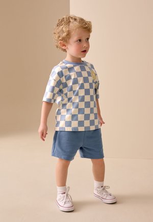 Malo dijete s kovrčavom kosom koje nosi plavo-bijelu kariranu košulju s motivom sunca, plave kratke hlače, bijele čarape i bijele tenisice, stoji.