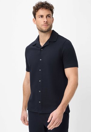 Mann mit kurzen lockigen Haaren, der ein schwarzes kurzärmeliges Hemd und passende schwarze Hose trägt und zur Seite schaut.