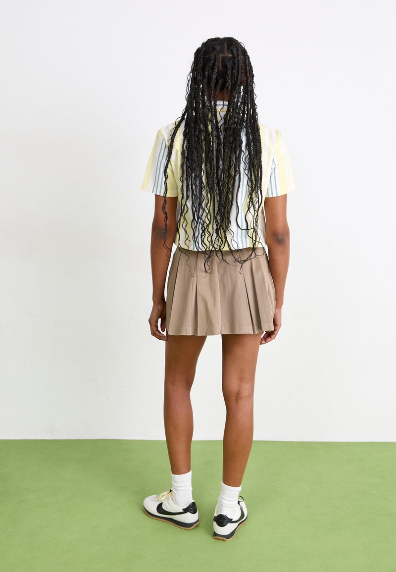 Camicetta a righe con maniche corte nei toni del giallo e blu chiaro, abbinata a una gonna plissettata color khaki, indossata con calze bianche e sneakers con dettagli neri.