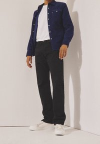 Camisa de pana azul con botones, camiseta blanca, jeans negros y zapatillas blancas. La camisa tiene bolsillos y botones plateados.