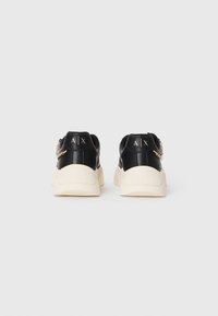 Baskets en cuir noir avec des semelles crème, arborant un logo doré sur le talon et un accent de chaîne décoratif sur le côté.