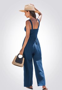 Navyblauwe jumpsuit van textuurstof, met wijde benen en verstelbare bandjes; gecombineerd met een rieten hoed en een kleine bicolor handtas.