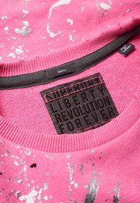 Rosa sweatshirt med en texturerad grafisk tryck och en svart etikett med texten "SUPERDRY LIBERTY REVOLUTION FOREVER." Storleksmärkningen säger "SMALL."