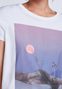 Camiseta de algodón blanca con un gran diseño impreso que presenta una luna rosa sobre un cielo de degradado púrpura y dunas de arena con vegetación escasa.