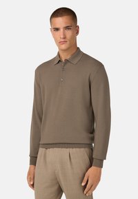 Khaki värvi pikkade varrukatega polo särk, millel on krae ja kolm nööpi, sujuva tekstuuriga ja soonikkudega mansetid, millele on sobitatud beežid püksid.