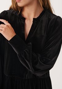 Abito di velluto nero con scollatura arricciata e maniche a sbuffo lunghe. Il tessuto presenta una texture liscia, che ne esalta l'aspetto elegante.