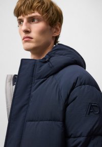 Granatowa kurtka puchowa z kapturem, z pikowanym wzorem, szarym wykończeniem wewnętrznym i haftowanym logo na rękawie.
