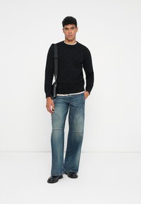Schwarzer Strickpullover mit strukturiertem Muster, kombiniert mit hellblauen Schlagjeans mit Nahtdetails und einer schwarzen Umhängetasche. Schwarze Schuhe.