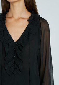 Schwarze Bluse mit V-Ausschnitt, ausgestattet mit einem Volantkragen und einer Knopfleiste vorne. Hergestellt aus leichtem, transparentem Stoff mit langen, fließenden Ärmeln.