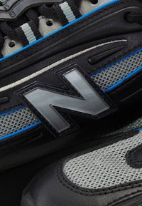Gros plan d'une basket New Balance noire et grise avec tissu en mesh, cuir noir, accents bleus et grand logo gris "N" sur le côté.