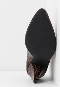 Zapato de tacón alto marrón texturizado con punta afilada y parte superior con patrón. Presenta una suela negra elegante y un tacón delgado y curvado.