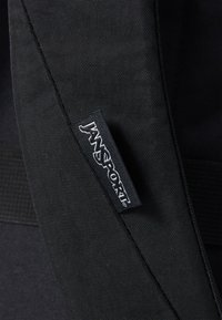 Zwarte stof met een gestructureerd oppervlak, waarop een zichtbaar Jansport-logo label is te zien. Beschikt over verstevigde randen en stevige stiksels.