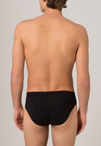 Slip da nuoto nero realizzato in tessuto liscio, con un design a vita bassa e una cintura semplice, visto da dietro su un torso nudo.