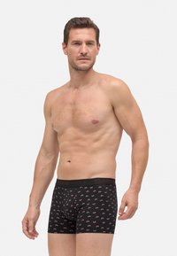 Hombre sin camiseta con cabello corto marrón, vistiendo calzoncillos negros con un patrón de bicicletas rojas y una cintura negra con el nombre de la marca.