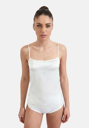 Woman with hair in a bun wearing a white satin camisole and matching white underwear, standing against a plain light background.