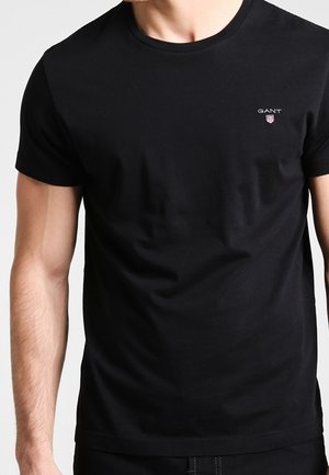 Homme portant un t-shirt noir uni à manches courtes avec un petit logo GANT sur la poitrine gauche.