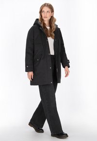 Sort parka-jakke med hætte, med en grå indvendig, frontlommer og knaplukninger, parret med sorte brede bukser og ankelstøvler.