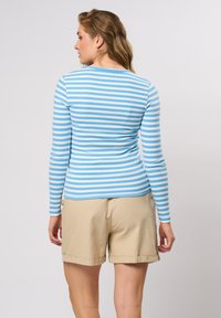 Langermet topp med lyseblå og hvite horisontale striper, laget av ribbestrikket stoff. Kombinert med beige shorts, vist fra ryggen.