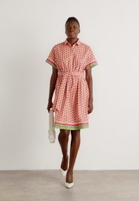 Kurzes Kleid in Orange mit einem kreisförmigen Muster, tailliertem Gürtel, grünen Akzenten und einem Saumdetail. Model kombiniert es mit einer weißen Handtasche.