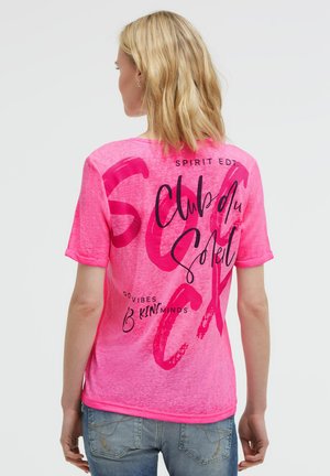 Young woman with blonde hair wearing a pink short-sleeve shirt with black and red text and blue jeans, shown from the back.