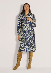 Frau trägt ein langärmliges, knielanges Kleid mit blau-beigem Blumenmuster, kombiniert mit beigen kniehohen Wildlederstiefeln.