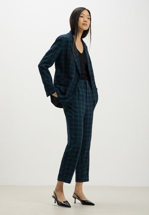 Blazer doppiopetto e pantaloni su misura in tessuto a quadri verde scuro. Top nero a scollo a V sotto. Tacchi neri a punta.