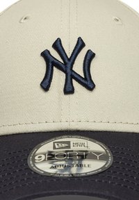 Gorra de béisbol ajustable con una corona beige, visera azul oscura y un logotipo bordado de la NY en azul marino en la parte frontal. Presenta una etiqueta reflectante de New Era.