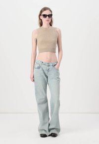 Un pull cropped beige côtelé, un jean bleu clair taille haute et des lunettes de soleil noires. Le mannequin se tient devant un fond blanc uni.
