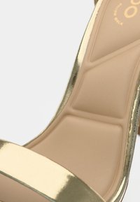 Sandale à talon haut métallique doré avec une semelle intérieure beige rembourrée. La chaussure présente une bride lisse et un design profilé pour le confort.