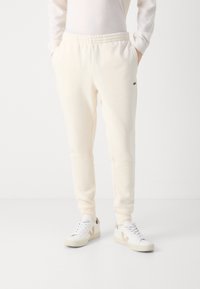 Crème-kleurige joggers met een elastische tailleband, zijzakken en een klein groen logo op de linkerheup, gecombineerd met witte sneakers.
