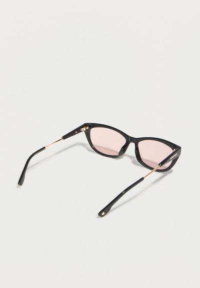 Γυναικεία γυαλιά ηλίου με μαύρο σκελετό τύπου cat-eye, λεπτά χρυσά μπράτσα και φακούς ανοιχτό ροζ απόχρωσης, τοποθετημένα σε ανοιχτόχρωμη επιφάνεια.