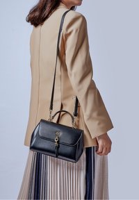 Kvinde iført beige blazer og plisseret nederdel, som holder en sort læderhåndtaske med skulderrem og guldlås.