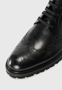 Stivaletto in pelle nera con finitura lucida, dotato di dettagli brogue, punta rotonda e lacci neri. Suola testurizzata per una buona aderenza.