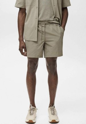 Chemise beige à boutons portée sur un T-shirt gris clair, assortie à des shorts beiges à cordon de serrage et des chaussures de sport claires. Tissu lisse, design décontracté.