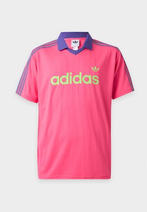Jeune homme aux cheveux tressés portant un t-shirt Adidas rose avec un col violet et des rayures, debout devant un fond blanc uni.