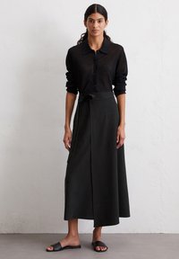 Camicia nera a maniche lunghe con bottoni, realizzata in tessuto leggero, abbinata a una gonna avvolgente scura, lunga fino al ginocchio, con cintura laterale e tasche.