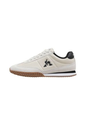 Zapatilla deportiva blanca con superposiciones de ante y sintético, acentos negros, logo característico y suela de goma texturizada.
