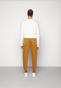Hanorac crem cu mâneci lungi, asortat cu pantaloni tapered din catifea în nuanță de maro. Încălțări sport albe completează ținuta. Fundal neutru fără alte detalii.