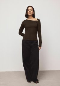 Brown long-sleeve top with an off-shoulder design and a gold accent. Paired with high-waisted black wide-leg trousers. Smooth texture.
