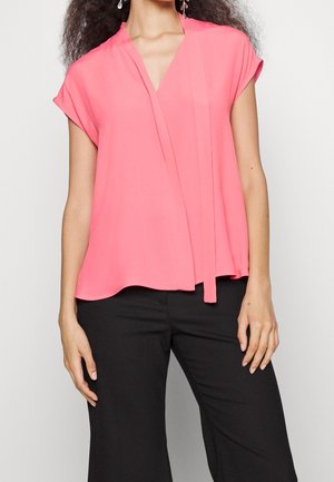 Kobieta ubrana w jasnoróżową bluzkę z krótkim rękawem i dekoltem w serek oraz czarne spodnie, stoi na tle neutralnego tła.